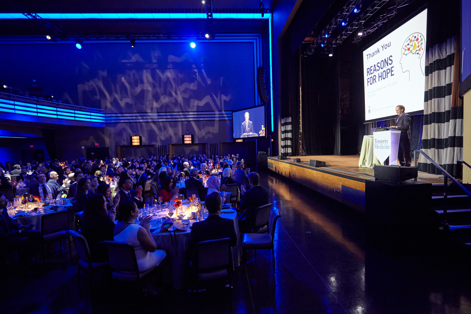 Host Rod Black entertains guests seated at dinner tables from the stage.